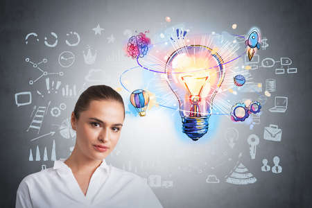 Portrait of confident attractive businesswoman in formal white tshirt. Colorful doodle cog wheels and lightbulb on concrete wall. Drawing with arrows, rocket, brain and network iconsの写真素材