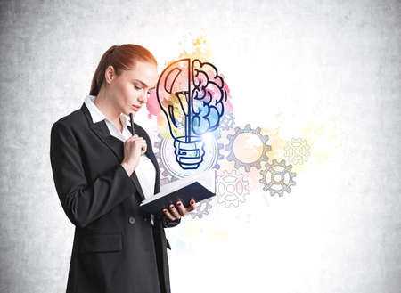 Office woman in black suit reading notebook with a pensive look. Gray concrete wall with colorful cogwheels and gears, colorful brain and light bulb doodle. Concept of start up and new ideasの写真素材