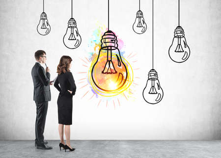 Businesswoman and businessman in office clothes, thinking near doodle light bulb drawn on concrete background, one colorful shining lamp. Concept of business idea and start upの写真素材
