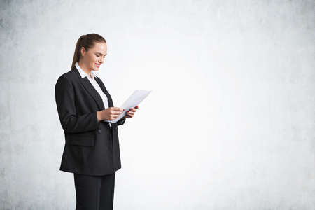 Office woman in black suit reading holding a business document in hands, copy space empty gray wall....