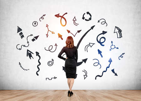 Woman in black dress, back view, with hand on waist. Confident person looking at different arrows with multiple direction on concrete wall. Concept of career development and challengeの写真素材