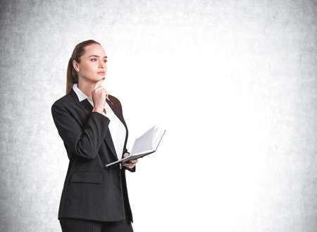 Attractive thoughtful young woman is holding a notebook and pondering how to optimize business process to boost sales of the company. Hand on chin. Concrete backgroundの写真素材