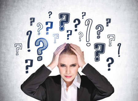 European attractive business woman holding her head by hands and pondering about career opportunities. Question marks drawn on concrete wall behind.の写真素材