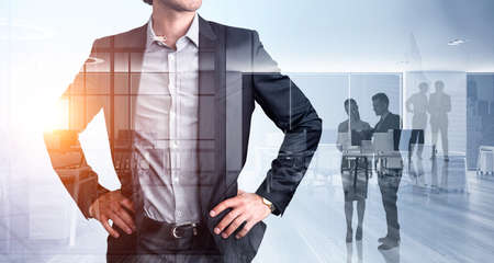 Confident corporate businessman, hands on waist pose. Diverse young professional business people aorking and looking for solutions in panoramic office space with skyscraper New York city view.の写真素材