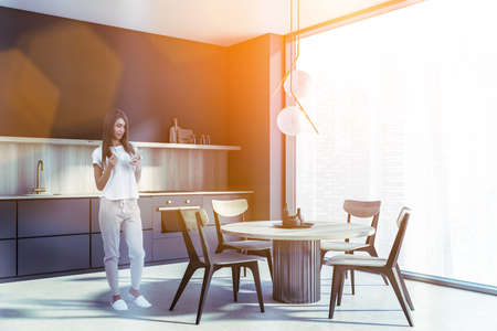 Woman is browsing the internet using phone and drinking coffee in modern kitchen room with dining...