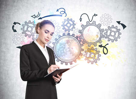 Young businesswoman in formal suit is taking notes into notebook, colorful gears sketches drawn on concrete wall behindの写真素材