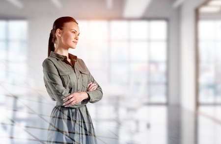 Confident attractive woman in casual hacky dress in crossed arm pose is pondering about life and education. Blurred panoramic office on backgroundの写真素材