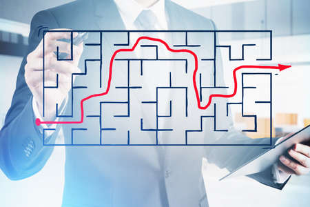 Businessman wearing formal suit is holding a pen and touching sketch with large labyrinth with red arrow showing the exit. Office in the background. Concept of thinking to find a better solutionの写真素材