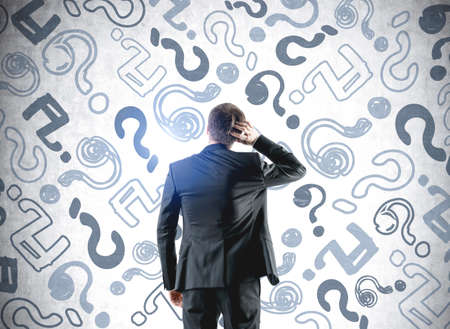 Thoughtful businessman wearing formal suit is holding head with his hand. Sketch with lots of question marks on chalkboard in the background. Concept of searching for solution to problemの写真素材