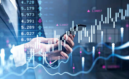 Businessman wearing formal suit is holding and touching smartphone with his finger. Blurred office workplace in the background. Financial chart and graph in the foreground. Concept of tradingの写真素材