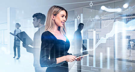 Smiling businesswoman wearing formal suit is holding clipboard. Group of business people work together taking notes for company. Office workplace in the background. Concept of teamwork and cooperationの写真素材