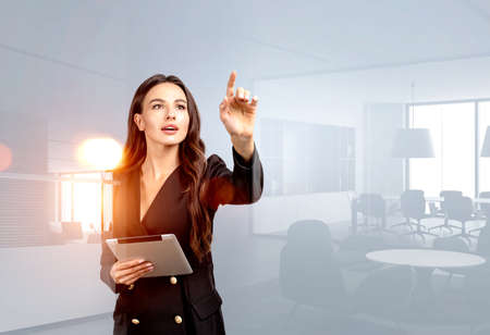 Thoughtful young attractive businesswoman wearing formal suit is standing and holding a tablet. Office workplace in the background. Concept of imagination and inspiration for creative ideasの写真素材