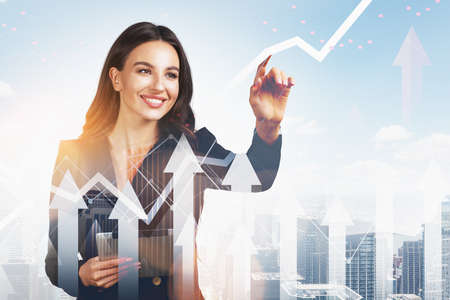 Smiling businesswoman wearing formal suit is touching screen with her finger. Financial graph and rising arrows in foreground. New York city skyscrapers in background. Concept of successful tradingの写真素材