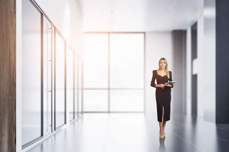 Smiling young attractive businesswoman wearing black dress is holding notebook and walking. Office workplace with panoramic window in the background. Concept of successful careerの写真素材