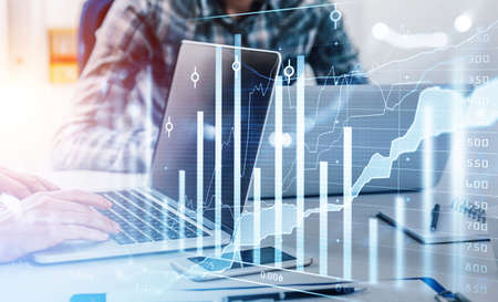 Two businessmen wearing tile shirts work together typing message on laptop. Office workplace with smartphone in background. Financial graph and chart, bar diagram in foreground. Concept of teamworkの写真素材