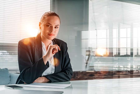 Smiling young attractive businesswoman wearing formal suit is touching her chin with pen. Office workplace with laptop and smartphone in the background. Concept of working processの写真素材