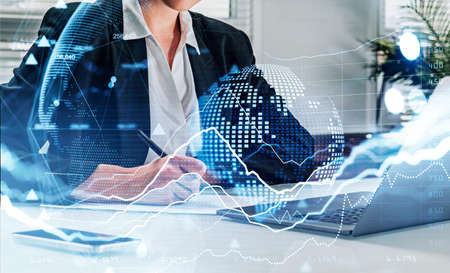 Businesswoman wearing formal suit is taking notes on paper. Office workplace with laptop and smartphone in the background. Financial chart, graph, virtual globe in the foreground. Concept of tradingの写真素材