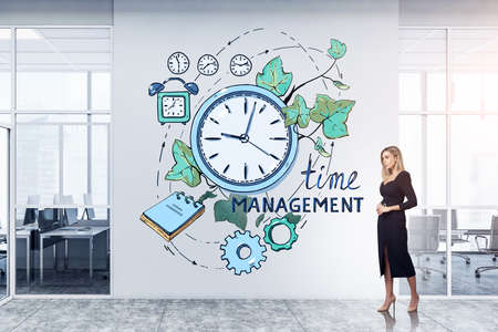 Young attractive businesswoman wearing black dress is standing near sketch with time management, leaves, notebook, clock, cogwheel on concrete wall at office workplace. Concept of creative ideaの写真素材