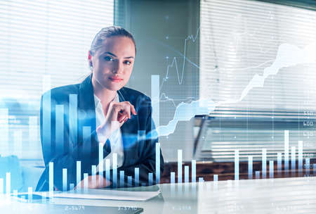 Businesswoman wearing formal suit is sitting and touching her chin with a pen. Office workplace with smartphone and laptop in the background. Financial graph and chart in the foregroundの写真素材
