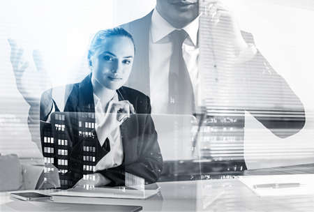 Smiling young attractive businesswoman wearing formal suit is sitting at office workplace with laptop in the background. Businessman is talking on smartphone. Concept of working processの写真素材