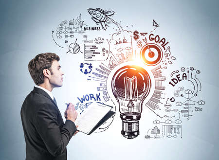 Businessman in formal suit takes notes and stands near sketch with light bulb, rocket launch, cogwheel, business, plan, teamwork on wall. Concept of imagination and inspiration for creative ideasの写真素材