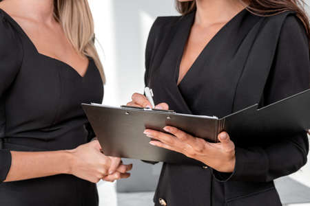 Businesswomen wearing formal suits are making contract for a profitable deal taking notes at clipboard. Office workplace with document in background. Concept of working process, teamwork, partnershipの写真素材