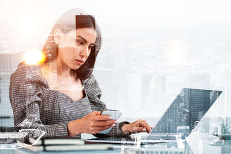 Office woman using phone, serious looking at the screen of laptop. Double exposure with New York...