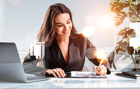 Office woman smiling dressed in black jacket, writing a business analysis. Technology and notebook on the table, double exposure with office room. Concept of business secretaryの写真素材