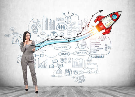 Office woman full length with device on parquet, doodle icons and colourful rocket with mind map on wall. Drawing with charts and network icons. Concept of product launchの写真素材