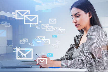 Businesswoman wearing gray dress is typing message on smartphone. Office workplace with desk, laptop and notebooks in the background. Concept of social media, communication and networkの写真素材