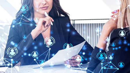 Young attractive businesswoman wearing formal suit is touching her chin with pen and talking to colleague. Office workplace in the background. Icons of social media in foreground. Concept of networkの写真素材