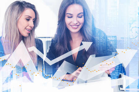 Two positive businesswomen in formal wears working together using tablet device. New York skylines on background. Digital financial charts with arrows on foreground. Double exposureの写真素材