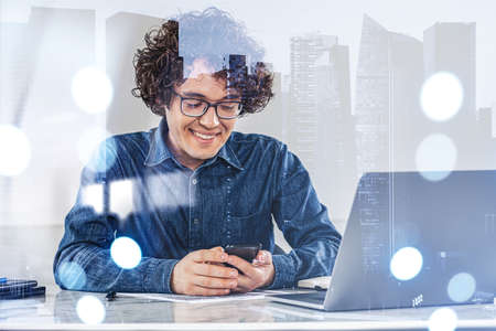 Business student man, happy look working with phone, laptop on the desk with papers. Double exposure of business city view. Concept of technologyの写真素材