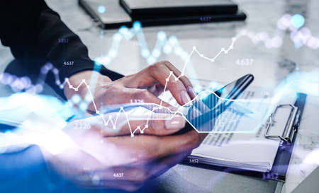 Businesswoman wearing formal suit is typing message on smartphone. Office workplace with clipboard in the background. Financial graph and chart in the foreground. Concept of trading and businessの写真素材