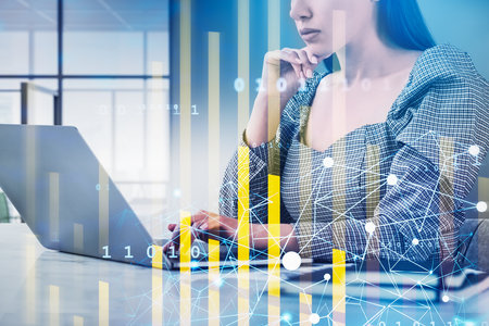 Businesswoman is typing message on laptop. Financial chart and graph with rising arrow and bar diagram in the foreground. Office workplace in background. Concept of trading and digital interfaceの写真素材
