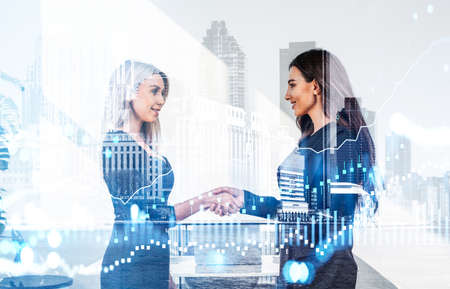 Two businesswomen wearing formal suits work together shaking hands after making profitable deal. Office workplace and New York city skyscraper in the background. Concept of teamwork and agreementの写真素材