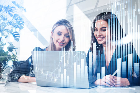 Two businesswomen wearing formal suits work together analyzing stock market on laptop. Office workplace in the background. Financial graph and bar diagram, forex candlestick. Concept of teamworkの写真素材