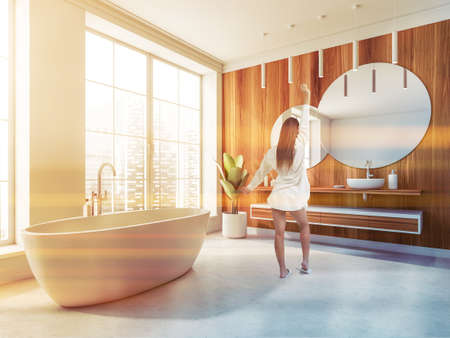 Young woman in the bathroom with tub and sinks, concrete floor and wooden wall with panoramic windows, toned image. Modern stylish room interior and person, lens flareの写真素材