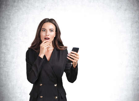 Shocked young attractive businesswoman wearing formal suit is holding smartphone in hands. Empty...