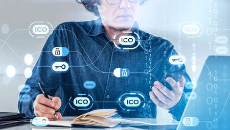 Office man take note and looking at the phone, laptop on desk. Initial coin offering hologram,...