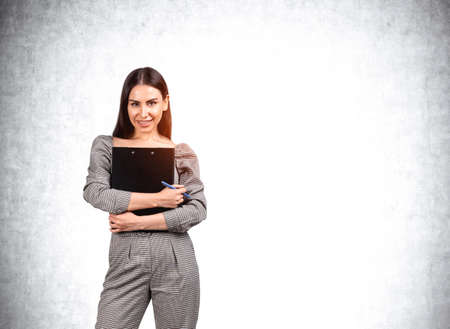 Smiling young attractive businesswoman wearing formal jumpsuit is holding clipboard in hands. Empty concrete wall in the background. Concept of imagination and inspiration for creative ideasの写真素材