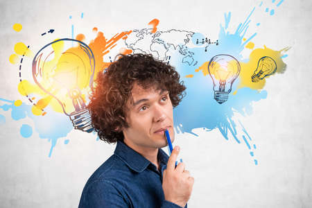 Thoughtful businessman wearing jeans shirt is touching chin with pen. Colorful sketch with three light bulbs, international map and human resources icon on concrete wall in the backgroundの写真素材