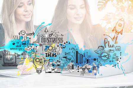 Two young attractive businesswomen work together typing on laptop and signing contract at office workplace. Sketch with rocket launch, start up, light bulb, bar and pie diagrams. Concept of teamworkの写真素材