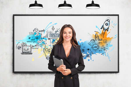 Young attractive businesswoman in formal suit is holding tablet near sketch with rocket launch, start up, laptop, light bulb, bar and pie diagrams, marketing on poster. Concept of creative ideasの写真素材