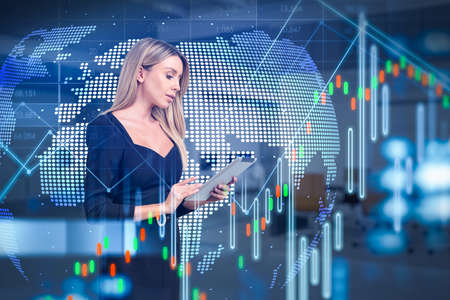 Young attractive businesswoman wearing formal dress is working on tablet. Blurred office workplace in the background. Financial graph and forex candlestick. Concept of communication via gadgetsの写真素材