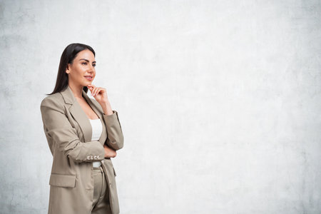Office woman in beige suit standing with inspired, thoughtful look. Boss smiling and making up plans and new ideas. Concept of startup and investment. Empty gray concrete wall, copy spaceの写真素材