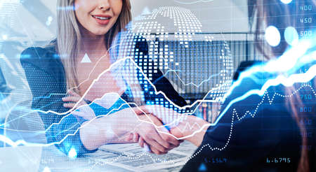 Two businesswomen wearing formal suits shakes hands after making deal. Office workplace with virtual globe and financial graph in the background. Concept of working process and agreementの写真素材