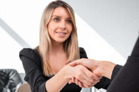 Smiling young attractive businesswoman wearing formal dress shakes hands with colleague. Office workplace in the background. Concept of teamwork, partnership, cooperation and coworkingの写真素材