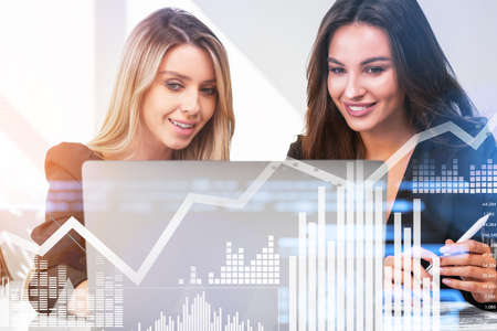 Businesswomen wearing formal suits are making contract for a profitable deal watching at laptop screen. Office workplace with document and bar diagram in background. Concept of trading on stock marketの写真素材