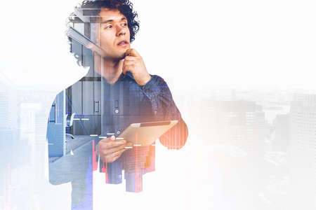 Pondering young handsome businessman wearing jeans shirt is holding tablet device. New York city skyscraper and office workplace in background. Concept of imagination and inspiration for creative ideaの写真素材